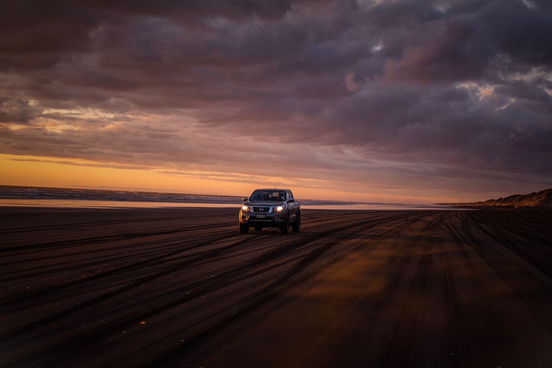 A 4x4 SUV driving on a sandy beach during a stunning sunset with dramatic clouds.