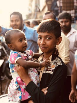 在繁华的户外市场，一个小男孩高兴地抱着一个女婴，画面捕捉了喜悦与家庭纽带