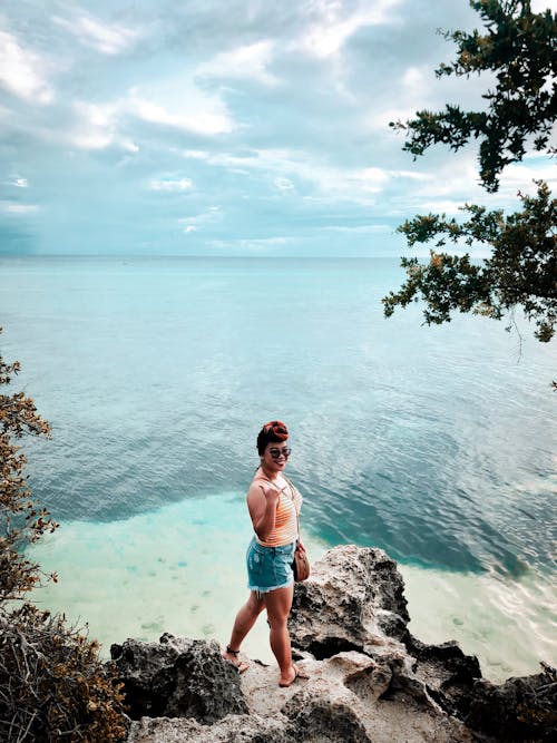 Photo of Smiling Woman in Blue Denim Shorts Standing on Seashore Rock Near Seashore