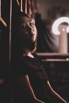 这是一幅沉思的肖像，描绘了一位年轻的亚洲女子在昏暗灯光下倚靠书架的情景