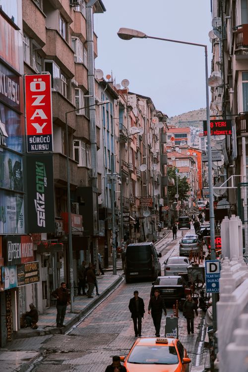Free Vibrant city street scene with people walking and cars on a busy urban road, lined with buildings. Stock Photo