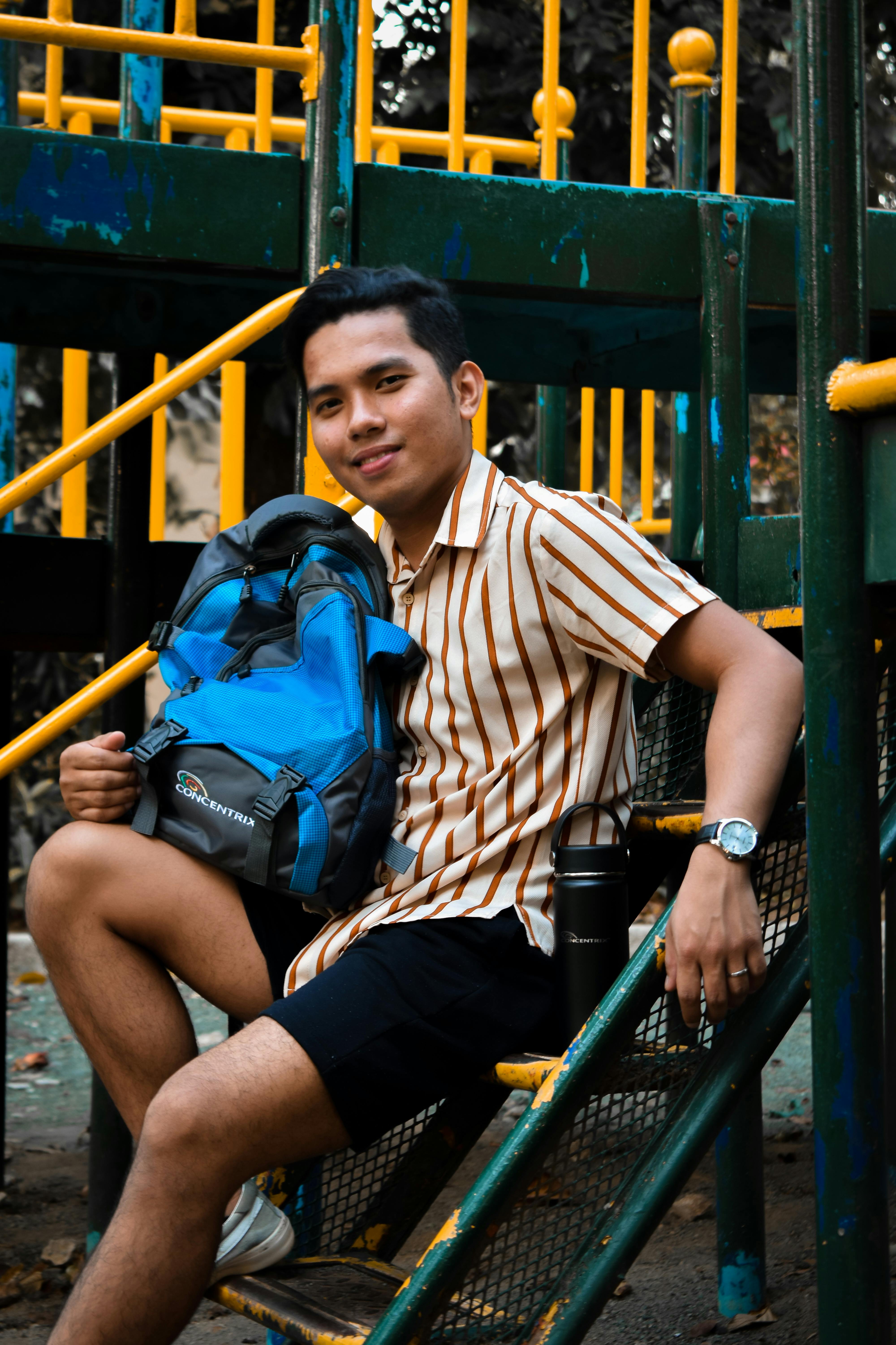 一个穿着随意的年轻人背着蓝色背包，在操场设备上微笑着