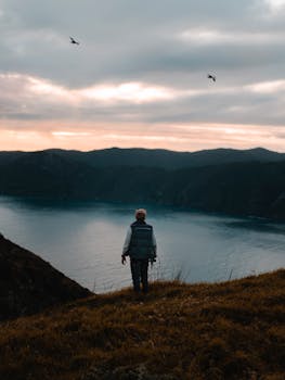 山上，一个孤独的人影俯瞰着宁静的湖泊，鸟儿在多云的天际飞翔