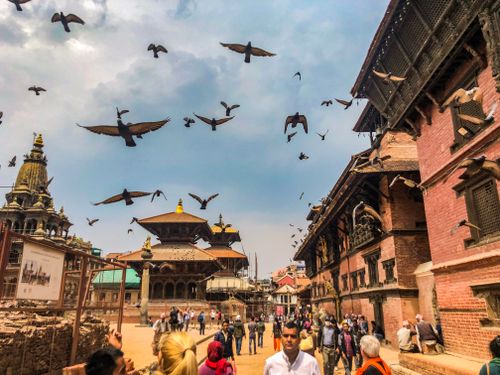 Free Vibrant scene at Patan Durbar Square with people, pigeons, and traditional Newari architecture under a cloudy sky. Stock Photo
