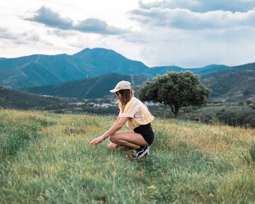 бесплатная Фотография женщины, стоящей на коленях на поле травы с горами на заднем плане, вид сбоку Стоковое фото