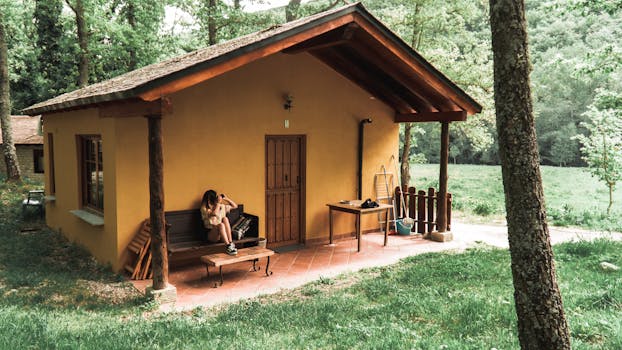 A woman sits on a bench outside a rustic cottage surrounded by trees and lush greenery.