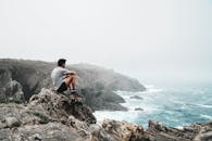 一个男人坐在悬崖边的岩石上，俯瞰着薄雾笼罩的海岸，陷入沉思