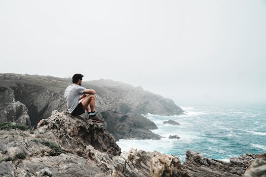 一个男人坐在悬崖边的岩石上，俯瞰着薄雾笼罩的海岸，陷入沉思