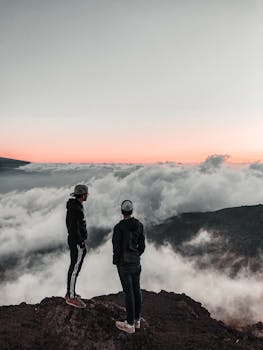 悬崖上，一个男子的剪影正欣赏着云彩与柔和的日出天空，体现了冒险与宁静