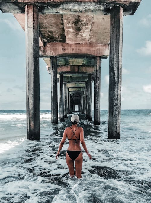 Free Back View Photo of Woman in Bikini Standing Below Wooden Oceanside Pier Stock Photo
