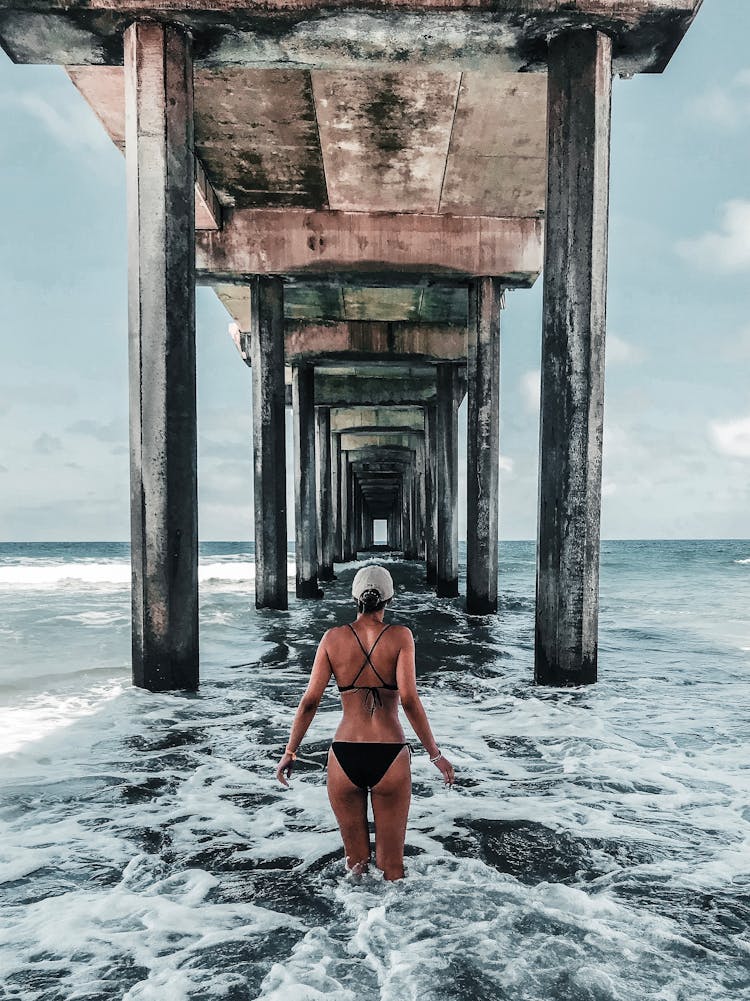 Back View Photo Of Woman In Bikini Standing Below Wooden Oceanside Pier