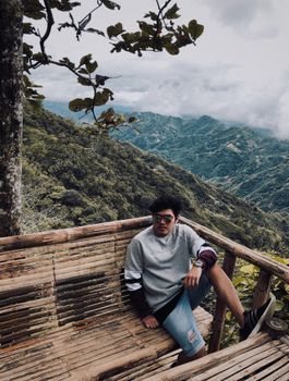 男子坐在木凳上，背后是山景，阴沉的天空平添了几分宁静