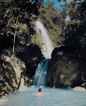 在宁静的瀑布旁，清澈的蓝色水中，一个人正在游泳，四周环绕着郁郁葱葱的绿色植物