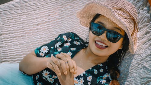 Close-up Photo of Smiling Woman in Black Floral Top, Sun Hat, and Sunglasses Lying on White Hammock 