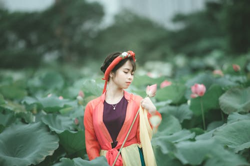 Selective Focus Photo of Woman in Colorful Outfit with Her Eyes Closed Holding Pink Flower Standing in the Middle of Flower Field