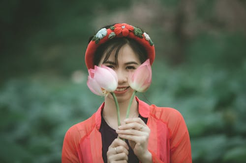 Selective Focus Photo of Smiling Woman Holding Up Pink-Lotus Flowers In Front Of Her Face