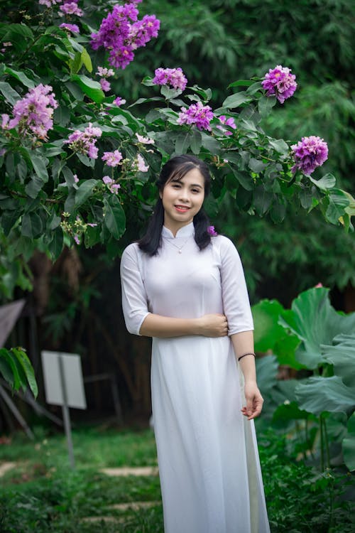 Foto Van Glimlachende Vrouw In Witte Jurk Die Zich Onder Paarse Bloemen Bevindt