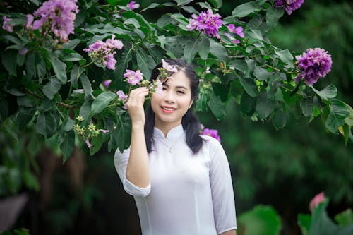 Portretfoto Van Glimlachende Vrouw In Witte Jurk Die Zich Onder Paarse Bloemen Bevindt