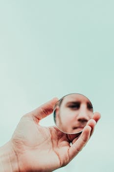 在柔和的天空背景下，一只手拿着一面圆镜，镜中映出一个男人的脸