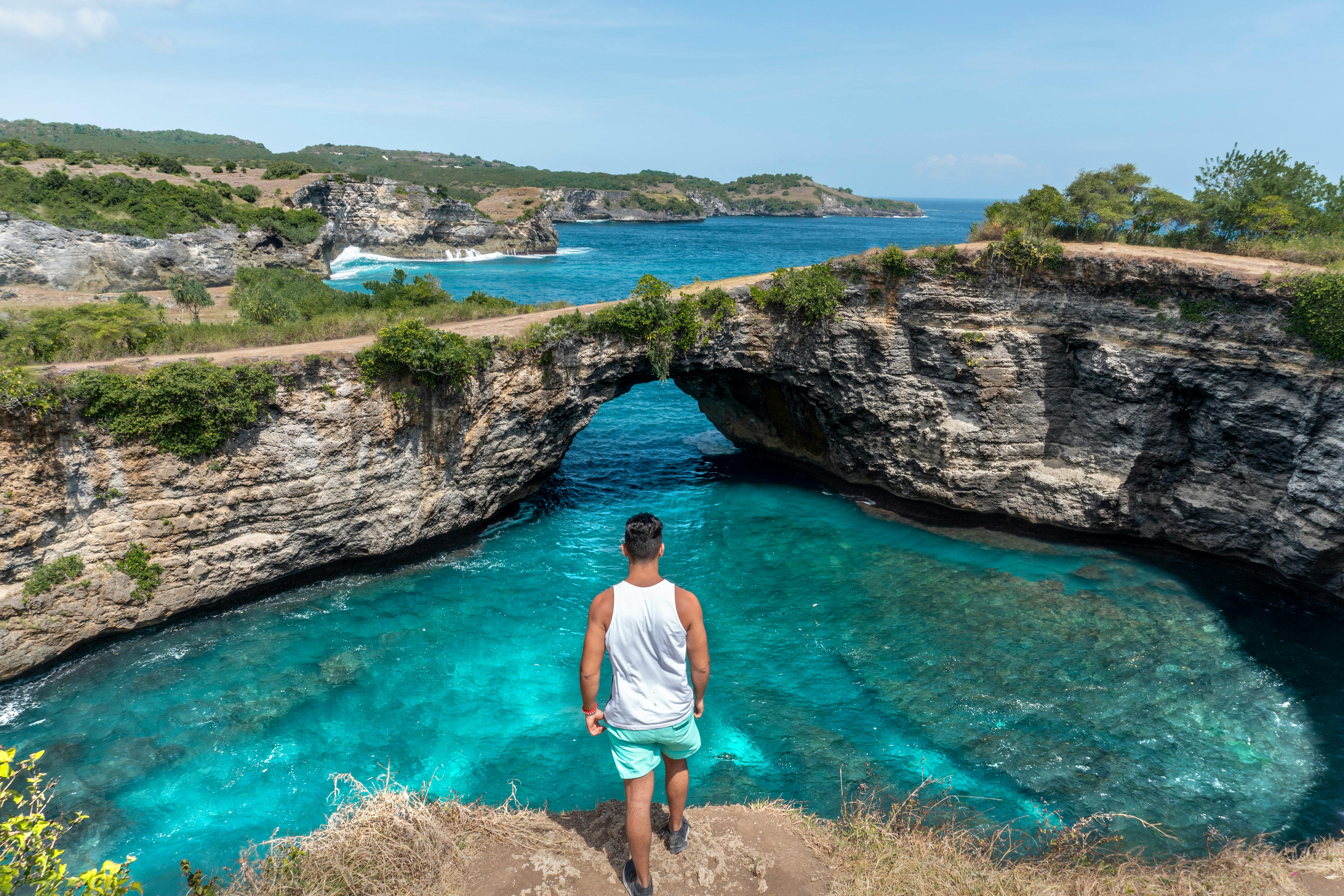 A man stands on a cliff's edge, taking in the stunning view of a tropical lagoon beneath a clear blue sky.