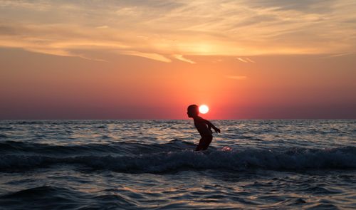 Free A child plays joyfully in the ocean waves as the sun sets, creating a stunning silhouette against the colorful sky. Stock Photo
