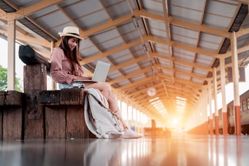 Foto De Baixo ângulo De Uma Mulher Sorridente Usando Um Laptop, Sentada Em Um Banco De Madeira Na Plataforma Da Estação Ferroviária
