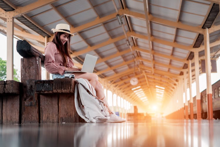 Low Angle Photo Of Smiling Woman Using A Laptop Sitting On Wooden Bench At Train Station Platform