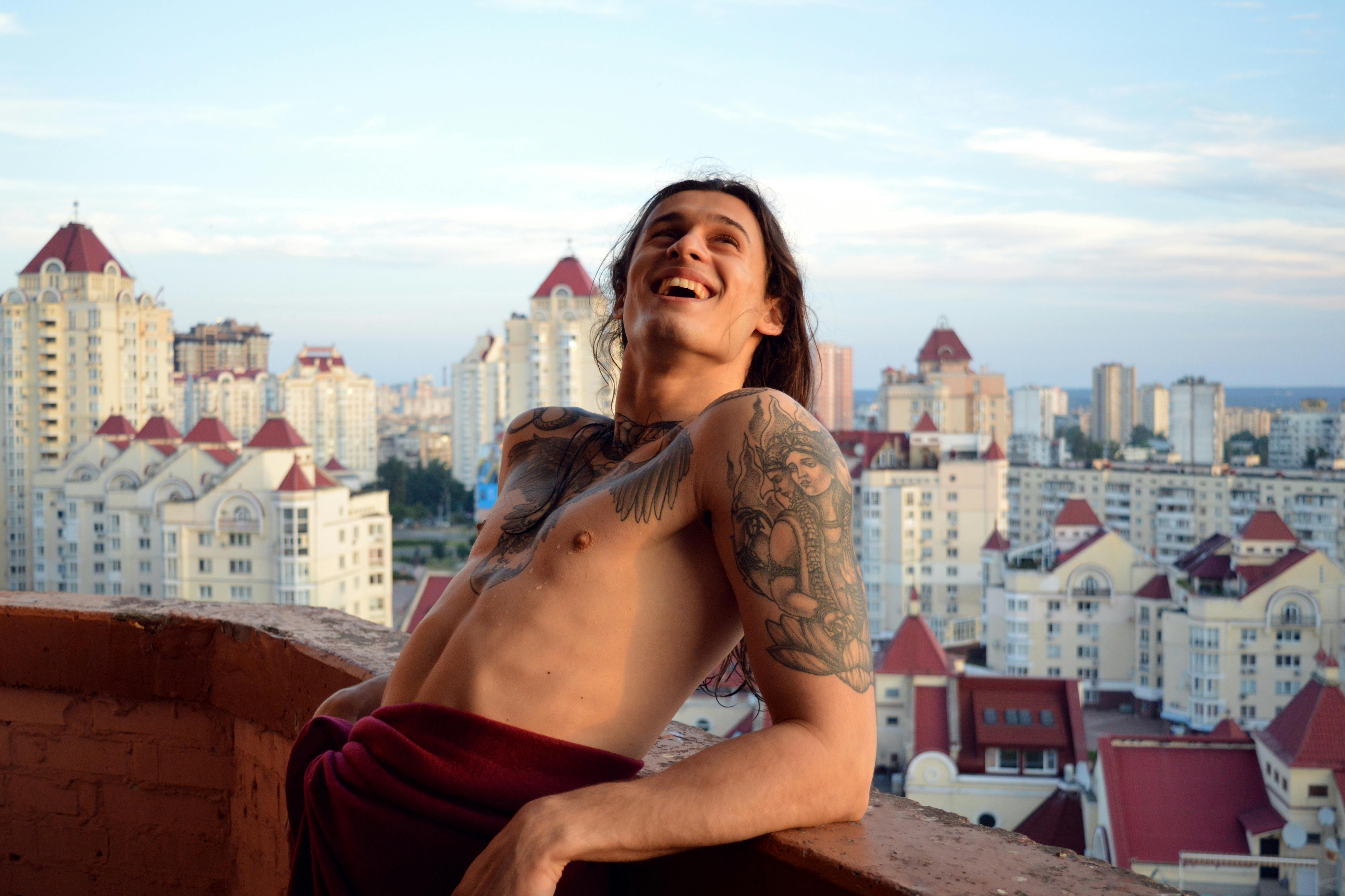 在晴朗的天空下，一位面带微笑的纹身男子正在阳台上欣赏城市景观