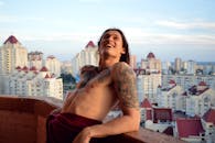 在晴朗的天空下，一位面带微笑的纹身男子正在阳台上欣赏城市景观