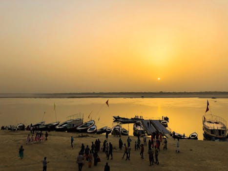 A serene sunset over a river with boats and people gathered at the shore, creating a picturesque scene.