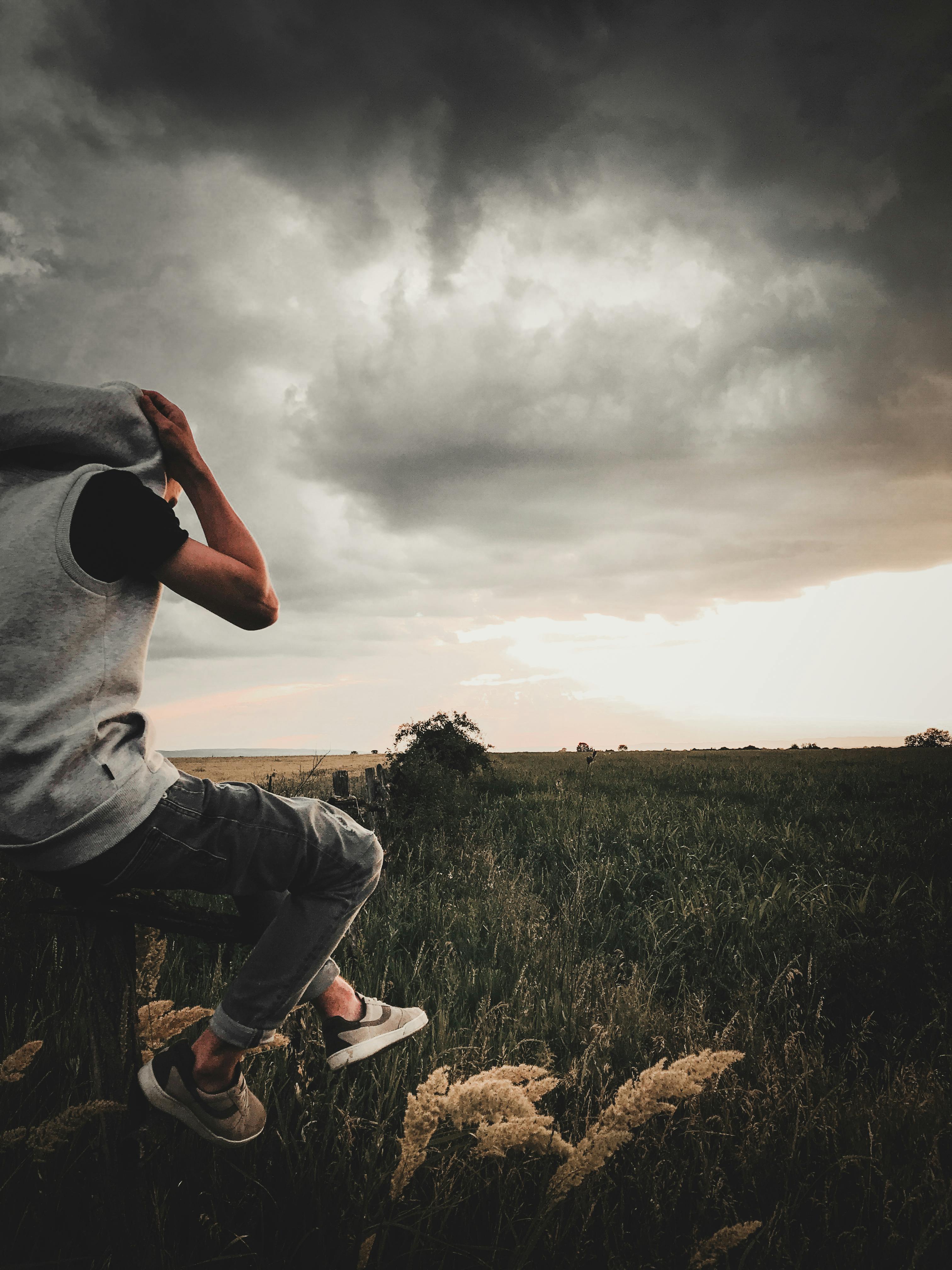 一个穿着休闲服的年轻人坐在暴风雨的天空下的草地上，这形象颇具戏剧性