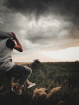一个穿着休闲服的年轻人坐在暴风雨的天空下的草地上，这形象颇具戏剧性