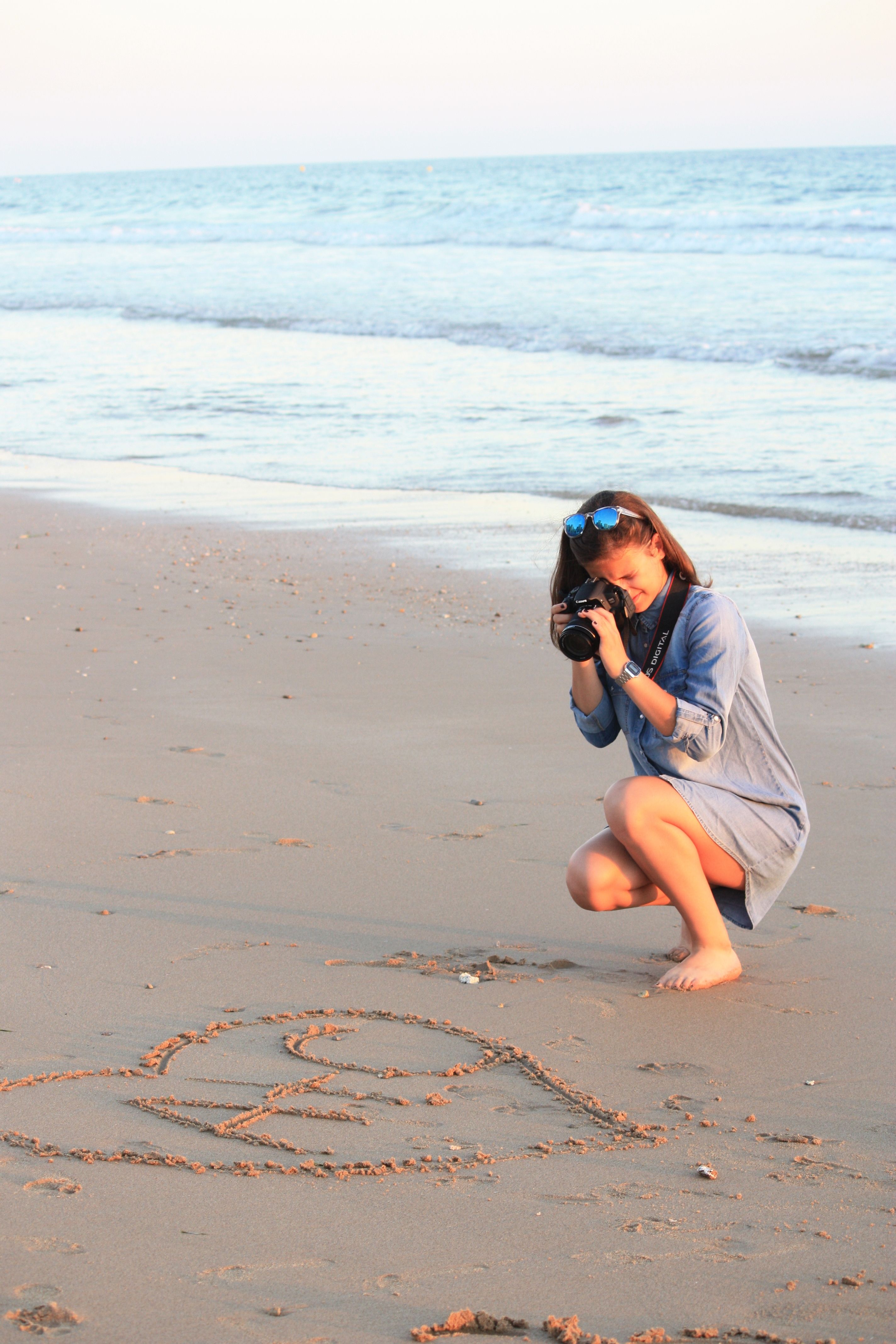 A woman takes photos on a sandy beach, capturing heart shapes in the sand.