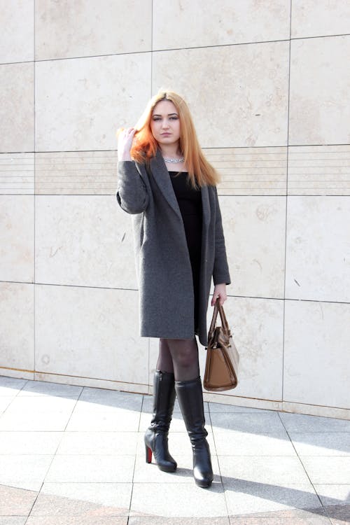 Foto De Mujer Con Abrigo Gris Posando Mientras Sostiene Su Cabello Y Lleva Una Bolsa De Cuero Marrón De Pie Frente A Un Muro De Hormigón Gris