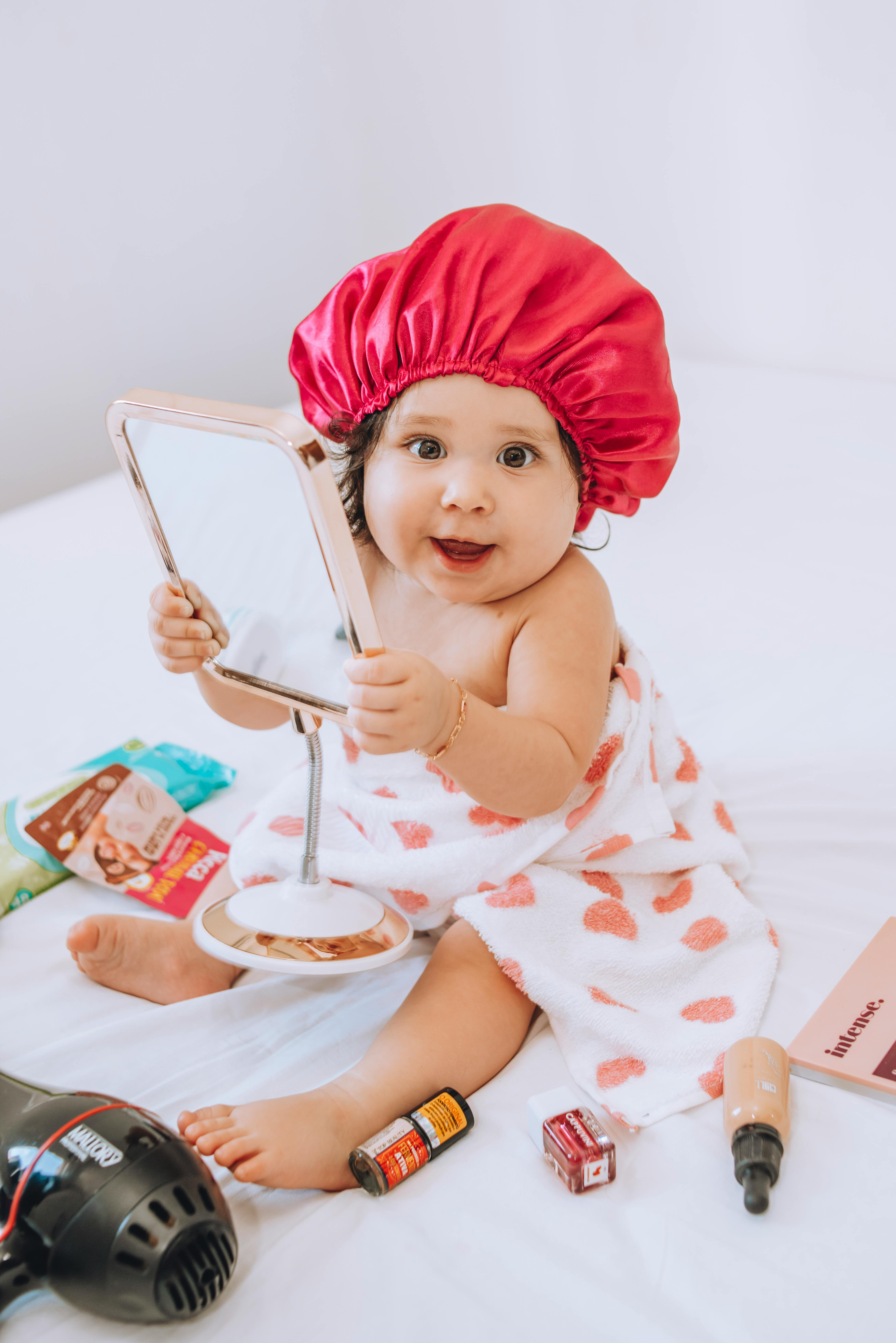 a little girl in a towel holding a mirror and sitting on a bed with makeup scattered around her