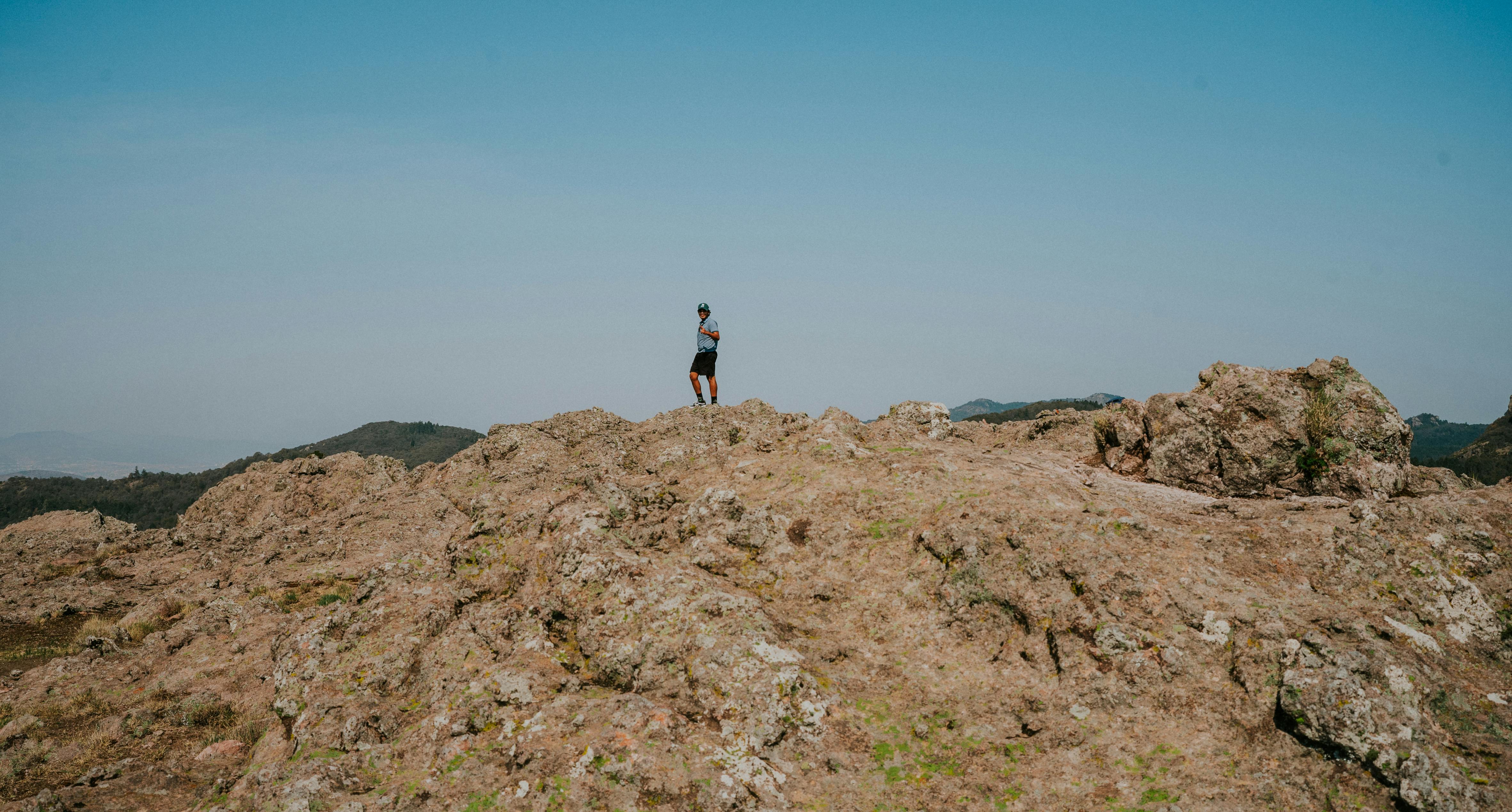 A solitary person stands on a rocky cliff, gazing at the vast landscape under a clear blue sky.