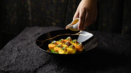 Free Close-up of a hand pouring sauce onto a gourmet dish in a dark, elegant setting. Stock Photo