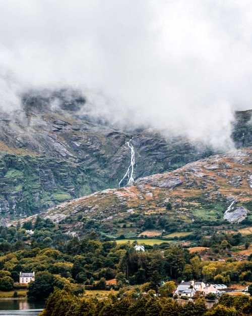 Free A stunning mist-covered highland waterfall with lush greenery and distant homes. Stock Photo