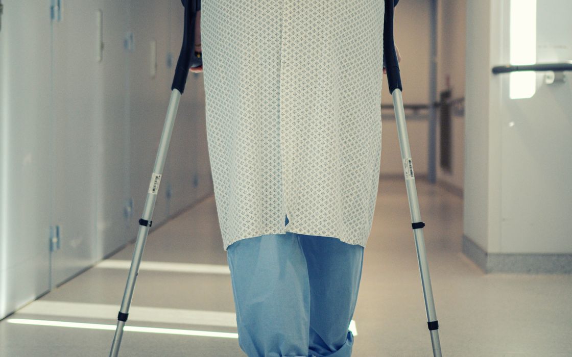 Patient in a hospital gown walking with crutches along a hospital corridor