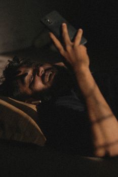 A man with a beard using a smartphone while lying indoors, smiling with earphones.