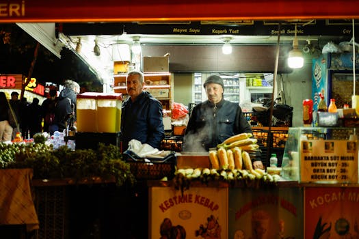 Two male vendors at a bustling street food stall at night, offering kebabs and corn.