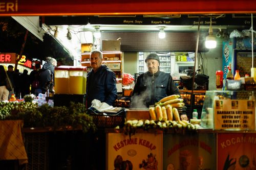 Free Two male vendors at a bustling street food stall at night, offering kebabs and corn. Stock Photo