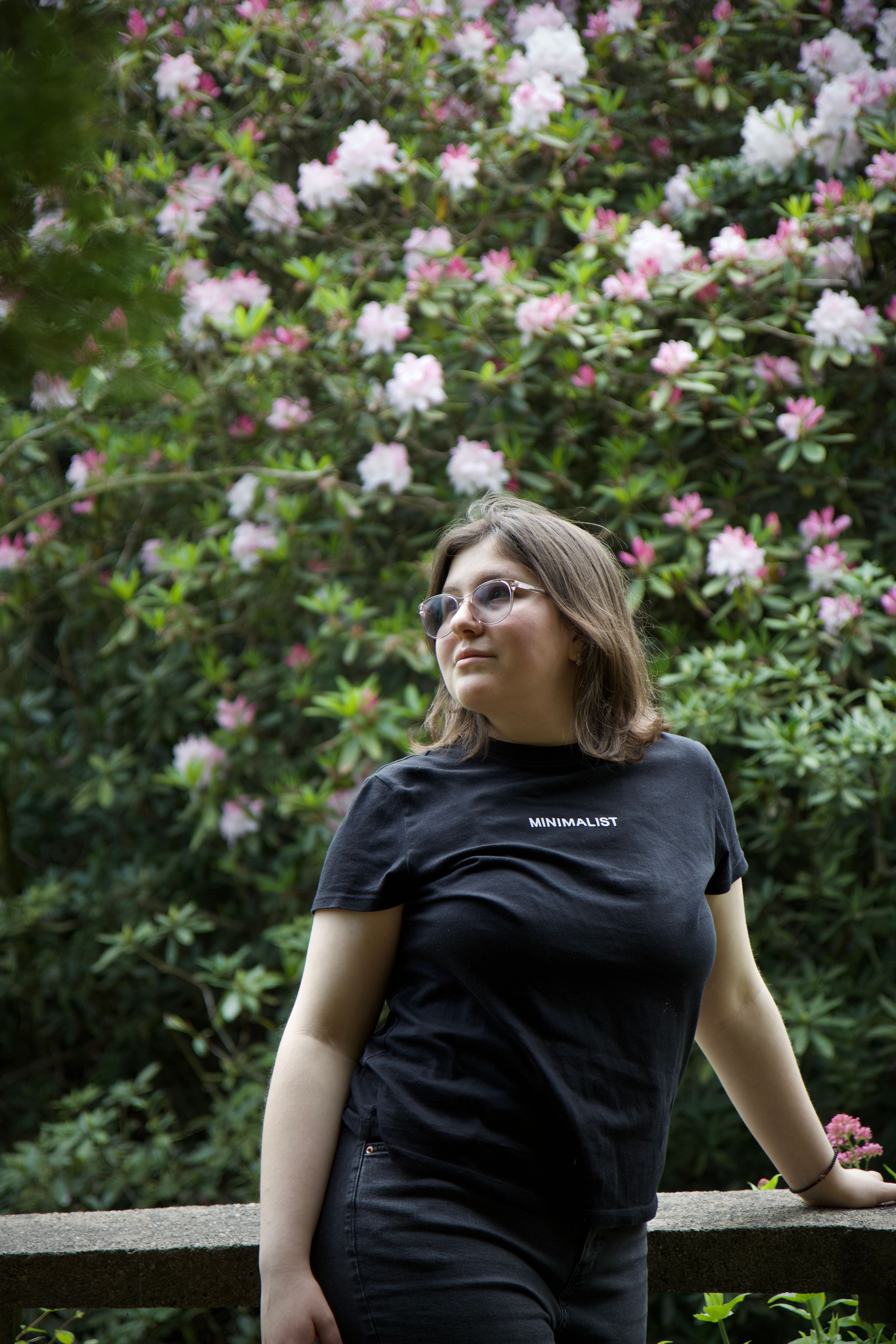 Woman with glasses in black t-shirt standing by railing in a garden of blooming flowers.