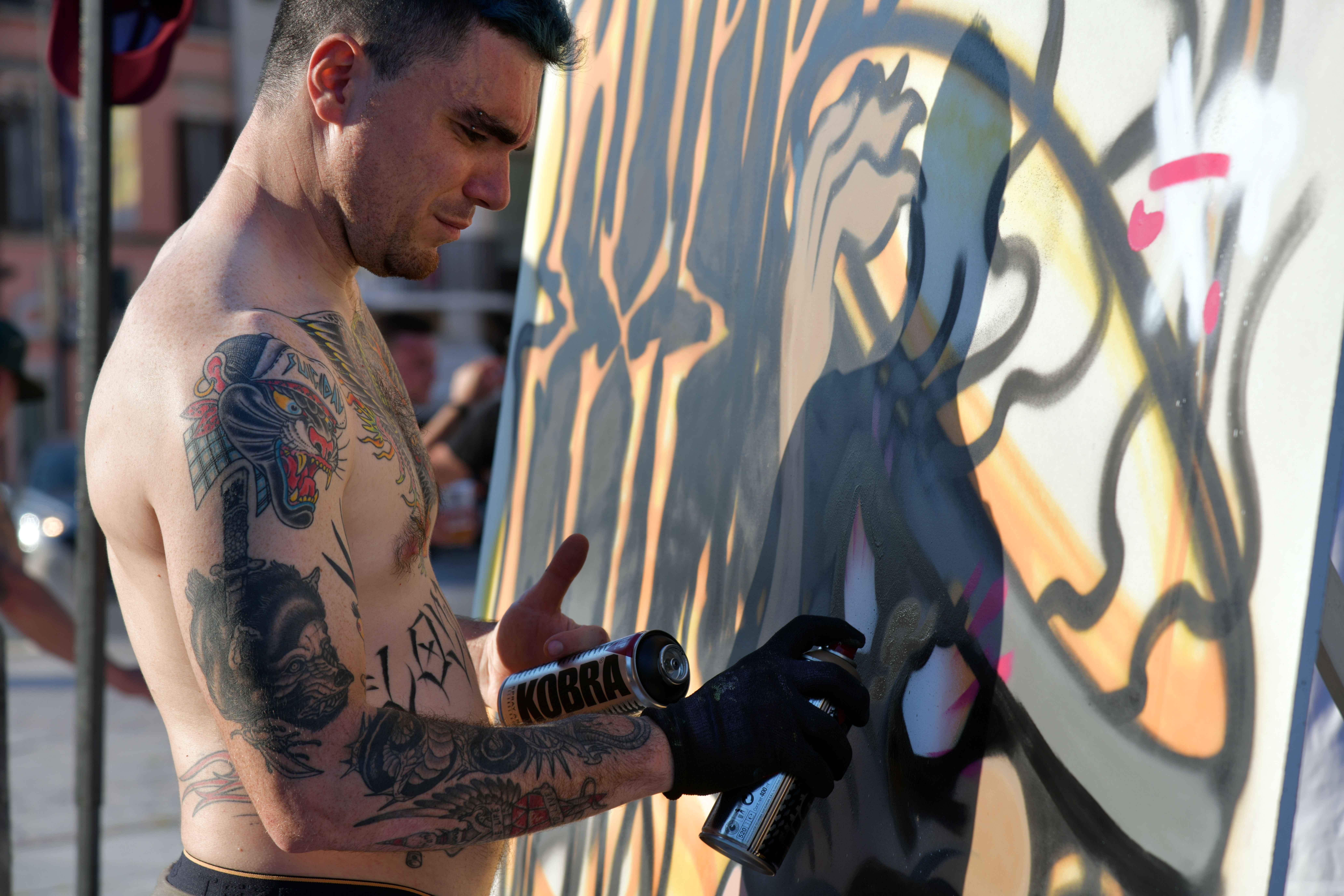 A tattooed street artist spray-paints a colorful graffiti mural on a sunny day in Villafranca di Verona.