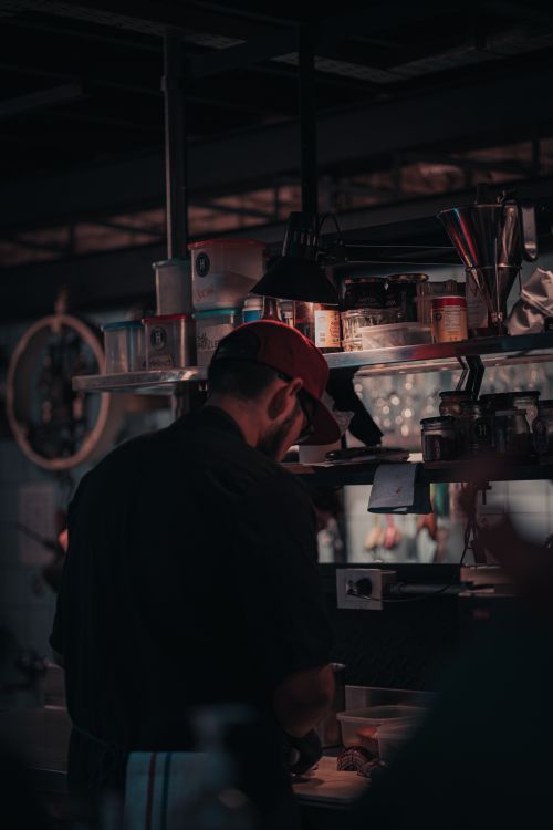 Free A barman wearing a cap works in a moody bar setting, surrounded by shelves of glasses and bottles. Stock Photo