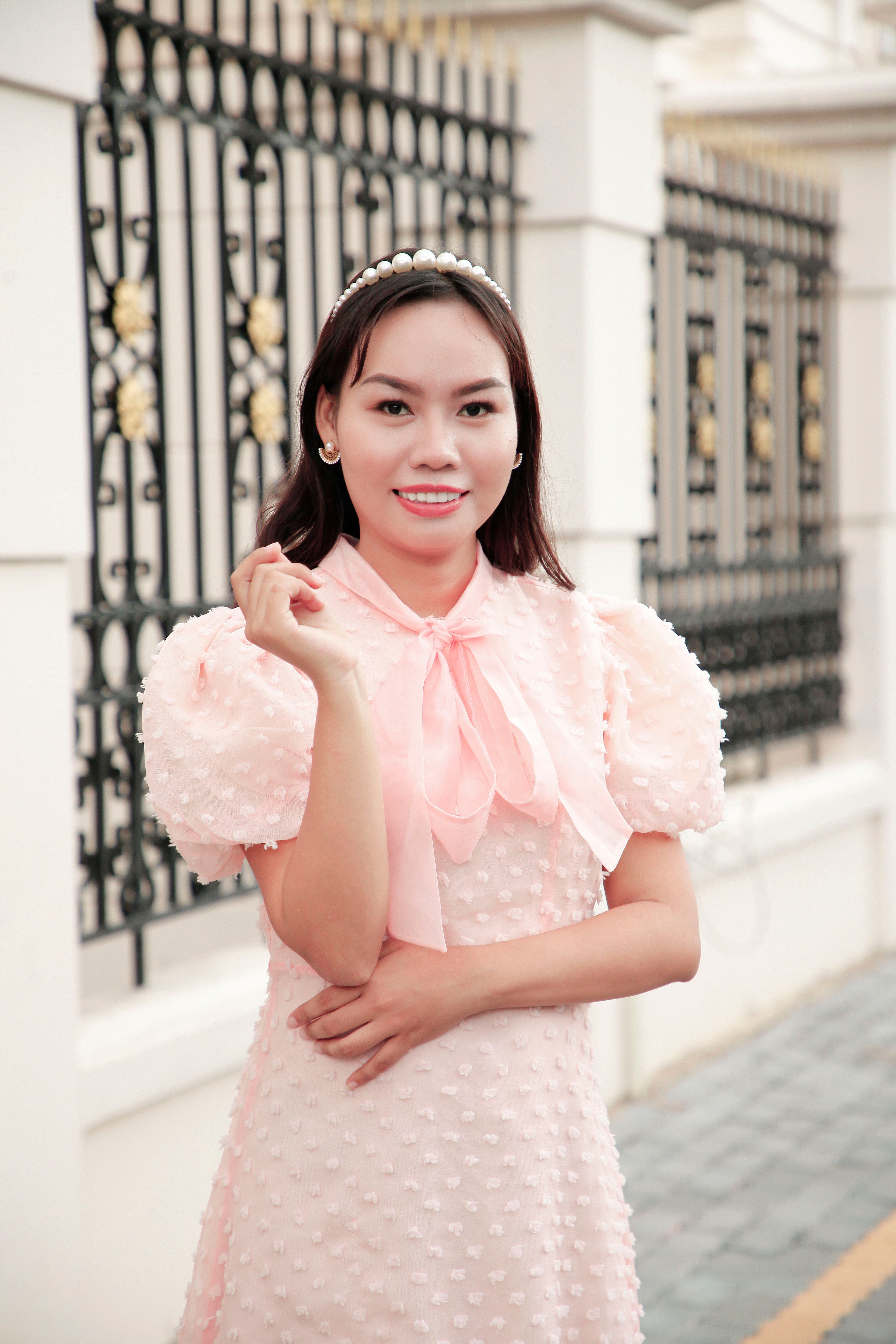 Elegant young woman posing in a pink dress by an ornate fence, urban setting.