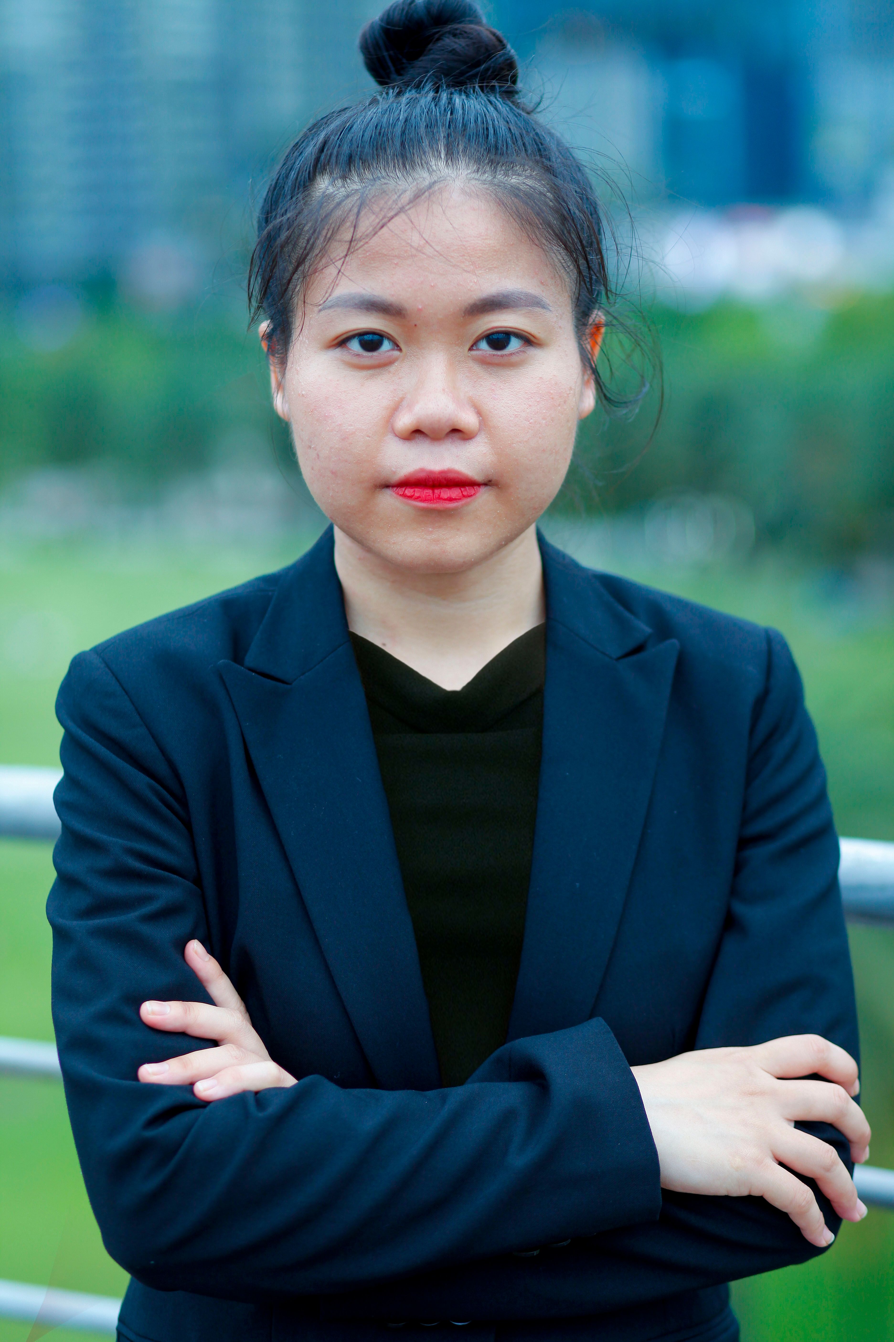 Young woman in black jacket posing confidently outdoors with cityscape in background.