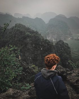 一名红发徒步旅行者站在岩石地形上，凝视着被薄雾吞没的山脉，展现出宁静的户外冒险氛围