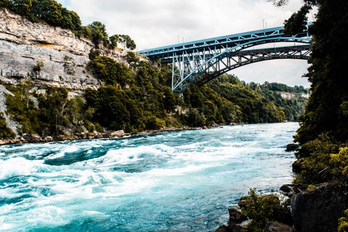 Free A scenic view of a river with a steel bridge, surrounded by lush green cliffs under a cloudy sky. Stock Photo