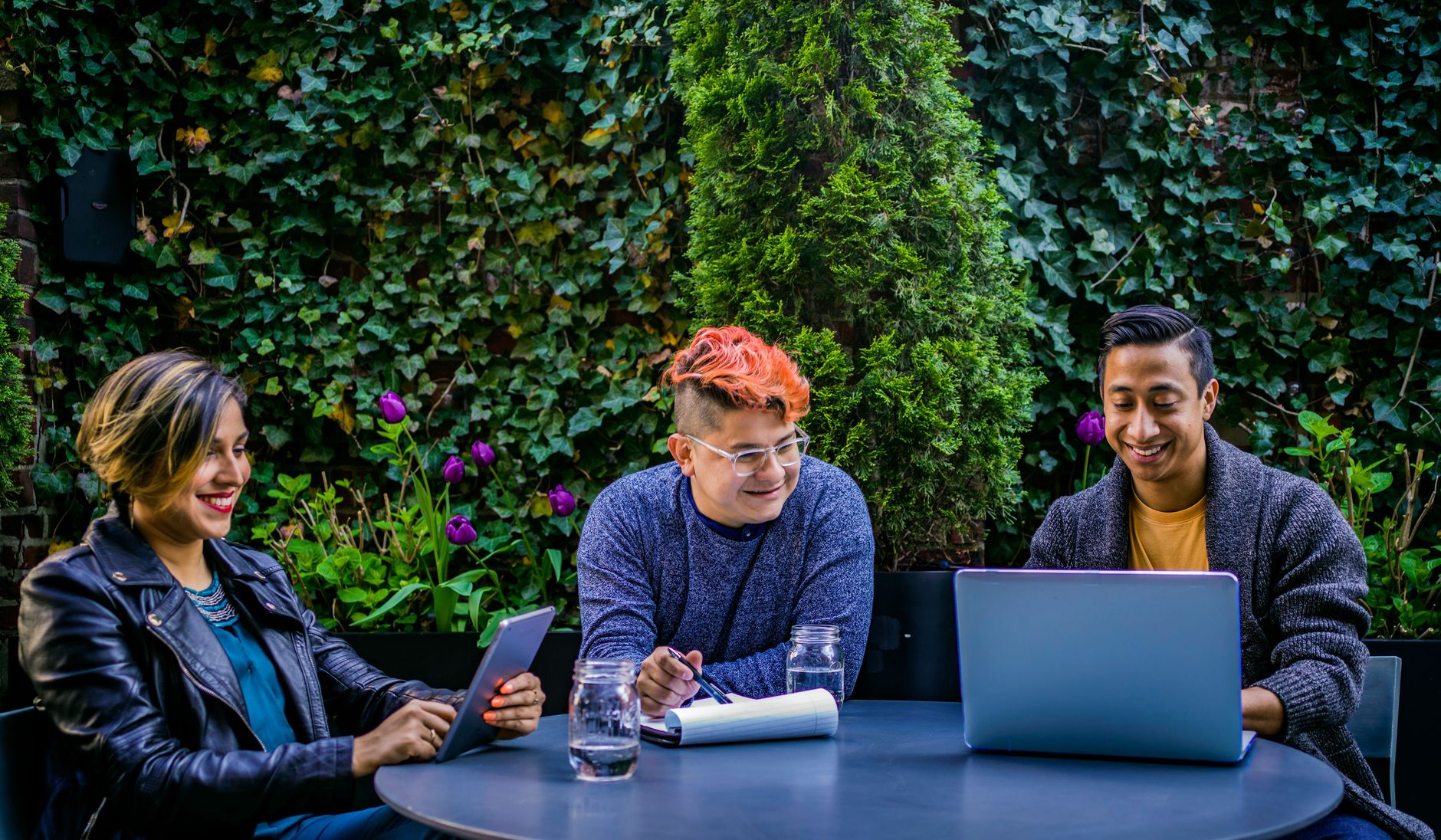 Three diverse adults engaging in an outdoor meeting with laptops and tablets in a garden.